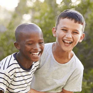 Two boys joyfully laughing and smiling together in a sunny park setting, surrounded by greenery and trees.
